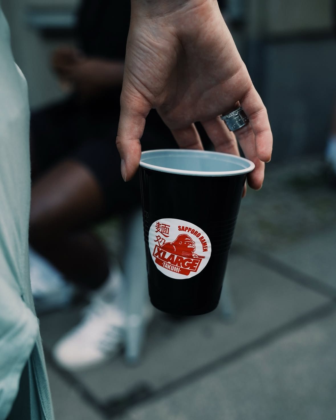 A hand holding a black cup with the XLARGE × Takumi Sapporo Ramen sticker